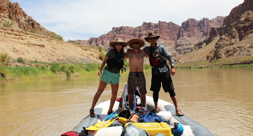 Three people embrace each other while standing on a raft. There are tall canyon walls framing the river in the background. 